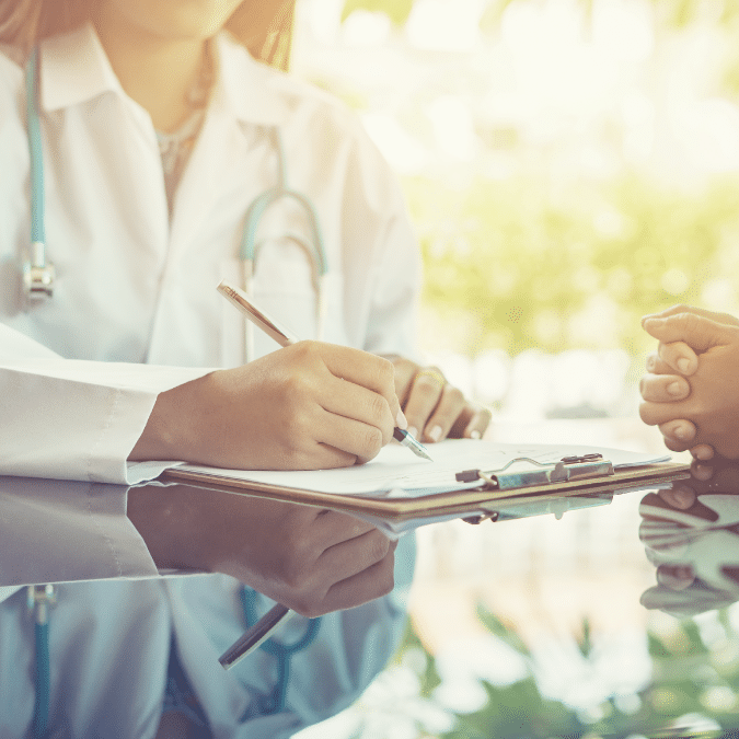 A doctor in a white coat writes on a clipboard while holding hands with a patient, offering comfort and support during a consultation at a medical office.