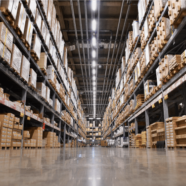 A wide-angle view of a large warehouse with tall shelves stacked with various boxes and packages, under bright overhead lighting, and a shiny polished floor.