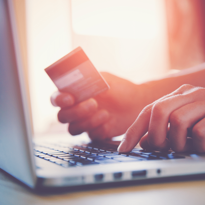 A person holds a credit card in one hand while typing on a laptop with the other, suggesting online shopping or electronic payment. The image is brightly lit with a soft background.