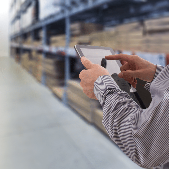 A person using a tablet in a warehouse, with shelves filled with boxes and products in the background—highlighting hands managing inventory through distribution center automation.