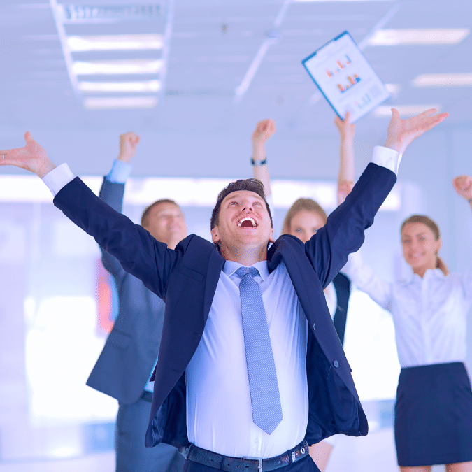 A group of businesspeople in formal attire celebrate enthusiastically in an office. One man stands at the front with arms raised and smiling broadly, while others cheer and raise their arms in the background.