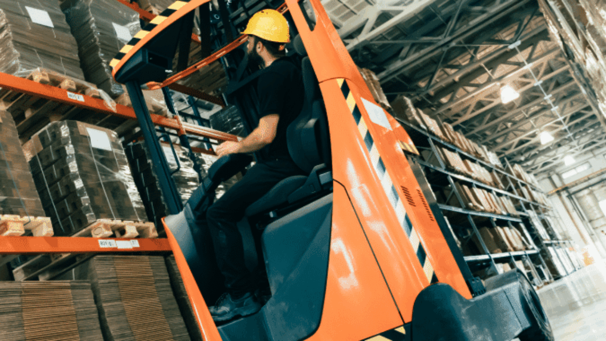 A worker in a yellow hard hat operates an orange forklift in a large warehouse, moving cardboard boxes stacked on tall shelves under bright lighting.