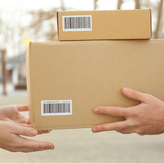 Two hands exchange two cardboard boxes with barcodes outdoors, one person handing the boxes to the other.