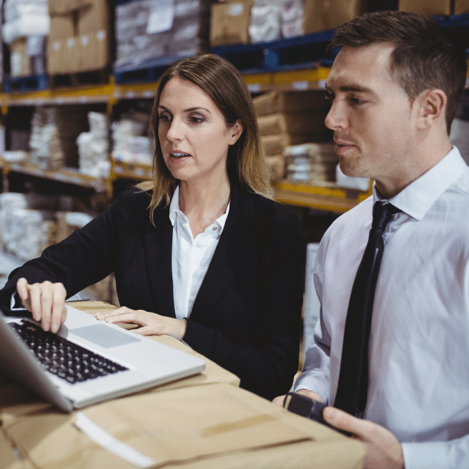 Two people in business attire look at a laptop in a warehouse, surrounded by shelves stacked with boxes and packages. One person is typing while the other observes attentively.