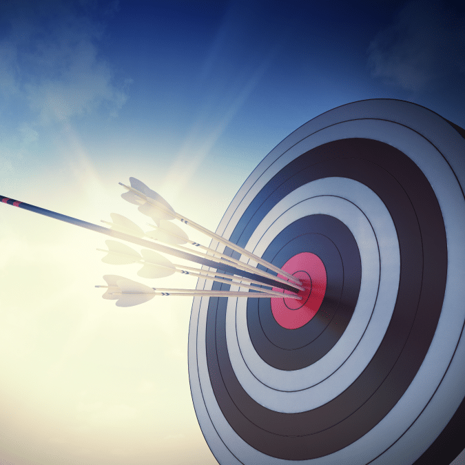 A close-up of a target with several arrows clustered in the bullseye, illuminated by sunlight against a blue sky, symbolizing accuracy and precision.