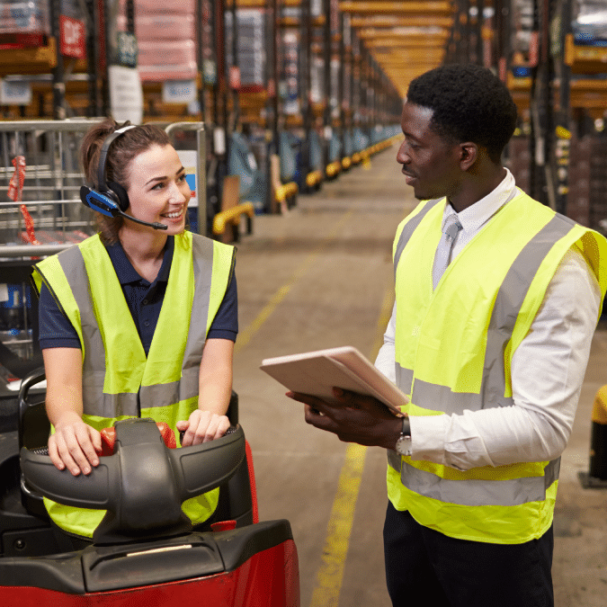 A woman wearing a headset and high-visibility vest sits on a forklift, smiling and talking to a man in a safety vest holding a tablet in a large warehouse.