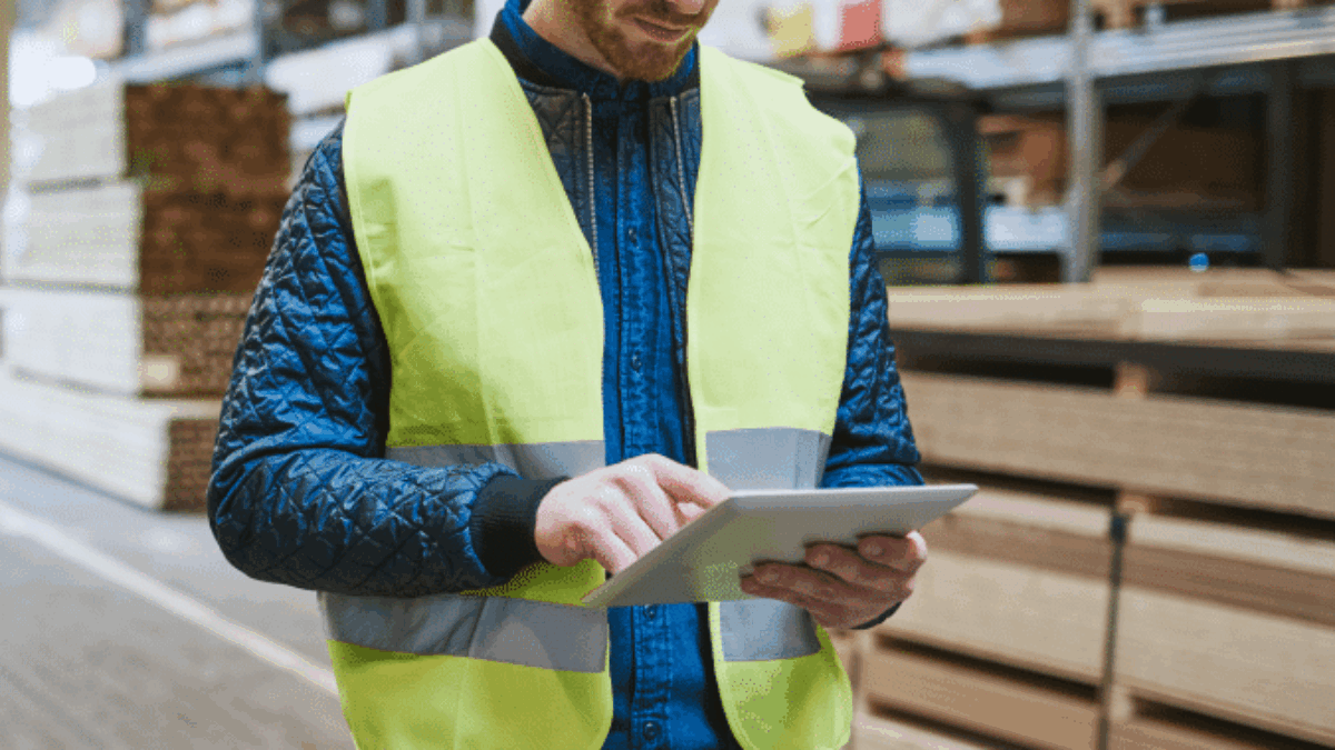 A warehouse worker in a yellow safety vest and tool belt uses a tablet to track tasks near stacked wooden boards, applying engineered labor standards in a large industrial storage area.