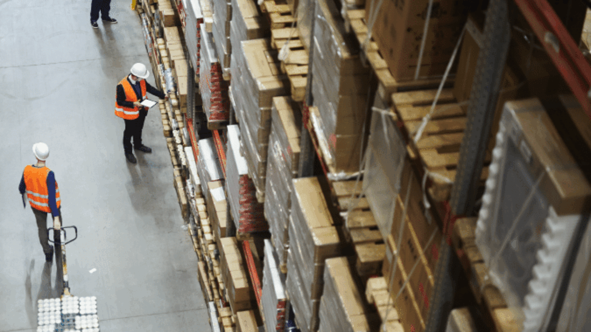 Three workers in safety vests and helmets are in a large warehouse with tall shelves stacked with boxes and pallets. One worker pulls a pallet jack while two others check inventory on clipboards.