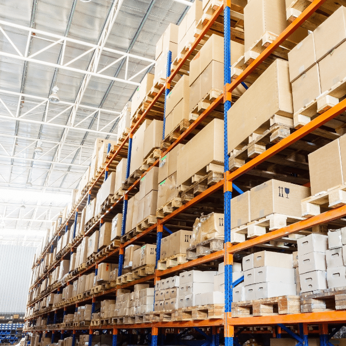Large warehouse interior with tall blue and orange metal shelves stacked with brown cardboard boxes and wooden pallets, extending into the distance under a high ceiling with exposed beams.