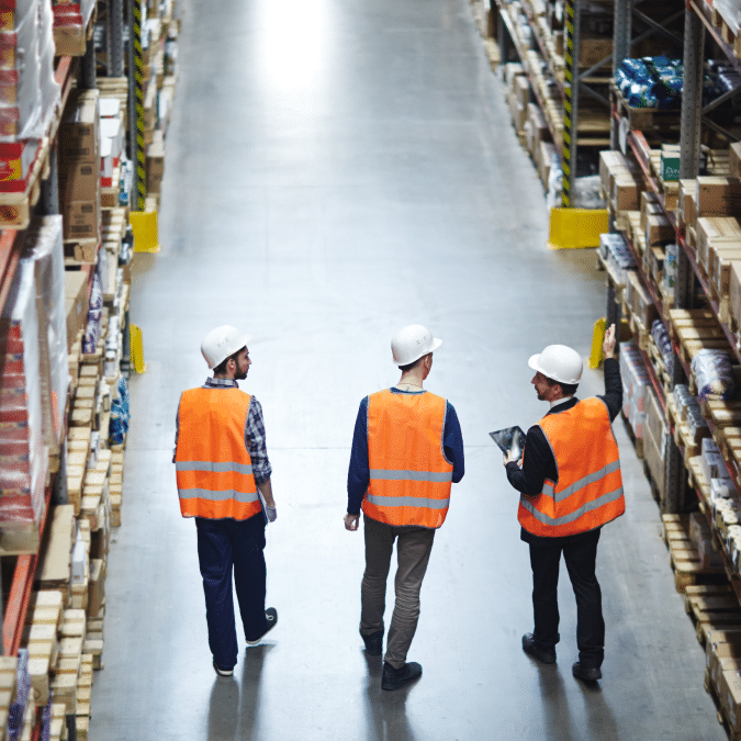 Three workers wearing orange safety vests and white hard hats walk down an aisle in a warehouse lined with shelves full of boxes and products. One worker gestures towards the shelves while another holds a clipboard.