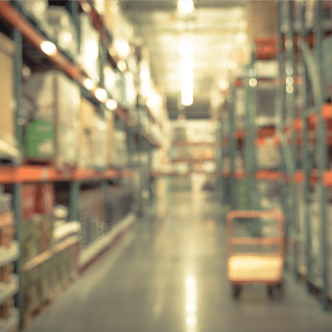 A blurred image of a warehouse aisle with shelves stacked with boxes and goods on both sides. A cart sits on the right side of the aisle under bright overhead lighting.