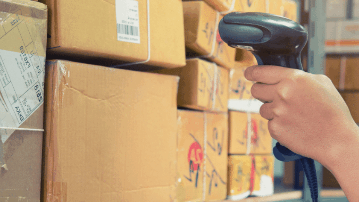 A person uses a barcode scanner to scan packages on metal shelves in a warehouse or storage area, surrounded by stacked cardboard boxes with shipping labels.
