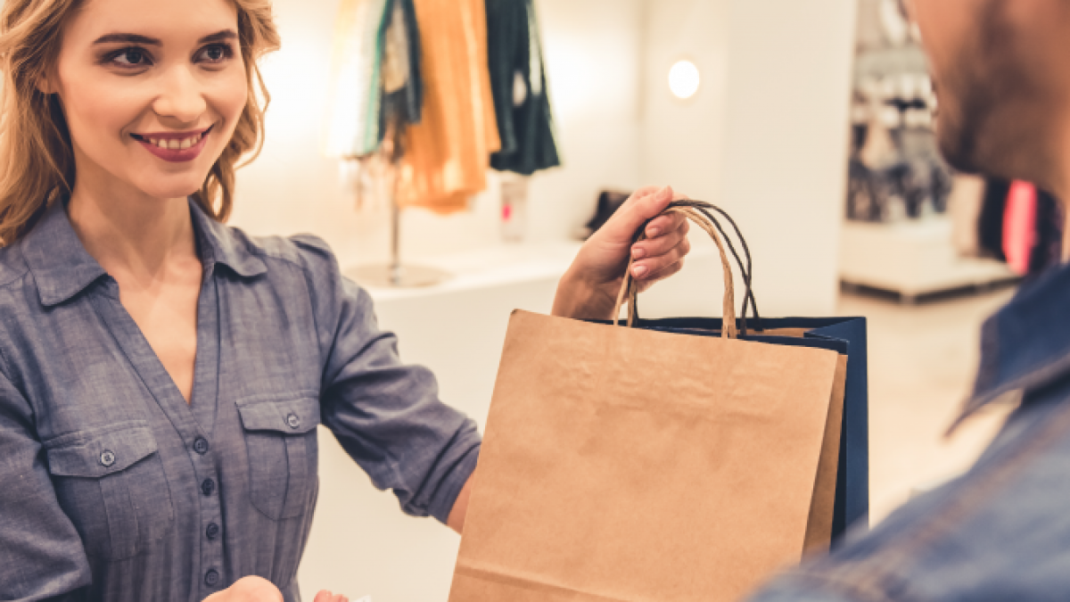 A woman at a retail counter smiles while handing a shopping bag to a customer, who is holding a credit card. Clothing and shoes are displayed on shelves in the background.
