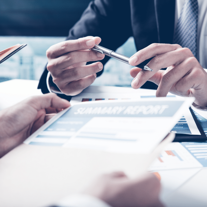 Two people in business attire discussing a summary report at a desk, with one person holding a pen and gesturing toward the document, suggesting a professional meeting or discussion.