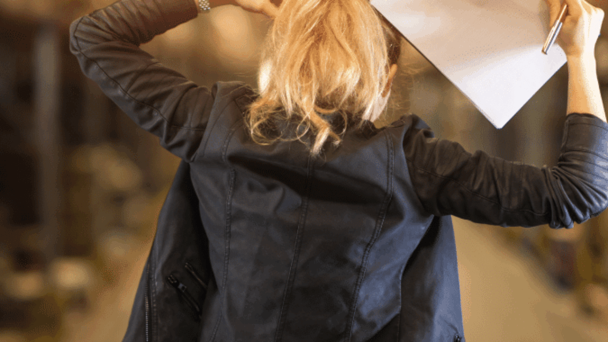 A woman with blonde hair, seen from behind, stands in a warehouse holding a clipboard and scratching her head, appearing thoughtful or puzzled.