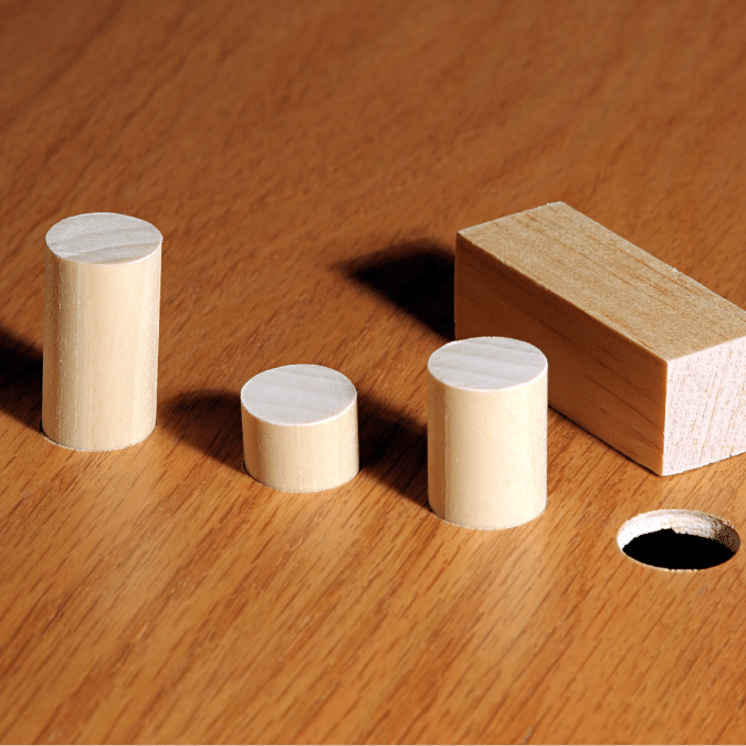 Three wooden cylinders and one wooden rectangular block sit on a wooden surface next to a round hole, illustrating the concept of shapes matching corresponding holes.