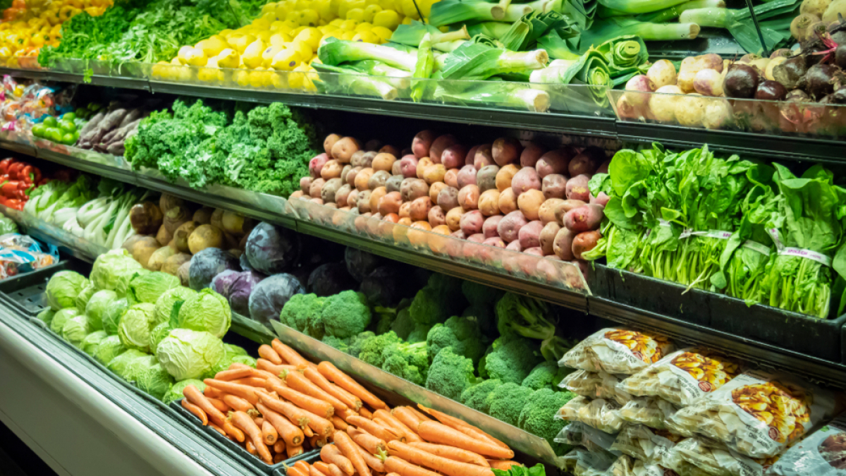 A grocery store produce section with neatly arranged vegetables including carrots, lettuce, broccoli, cabbage, potatoes, kale, squash, leeks, radishes, and packaged vegetables on multiple shelves.