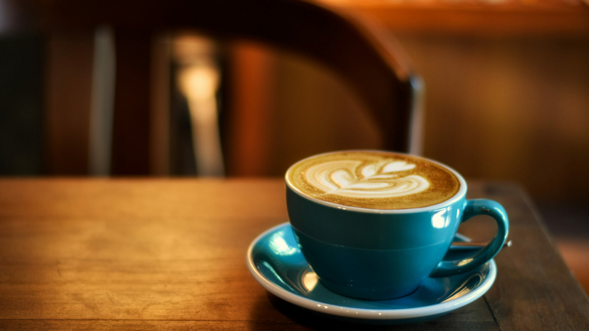 A blue cup and saucer with a latte featuring leaf-shaped latte art sits on a wooden table, capturing a cozy food and beverage moment against a blurred chair and warm-toned background.