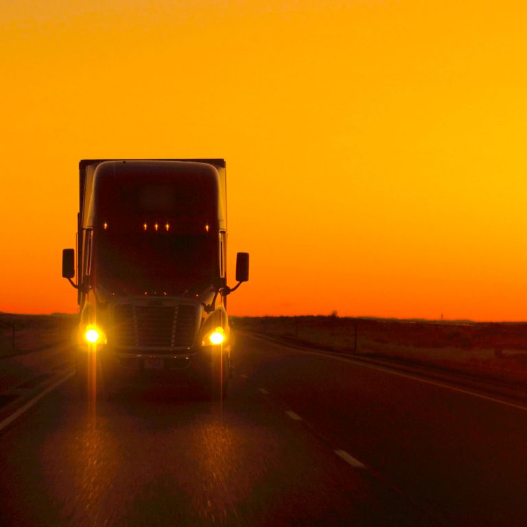 A large semi-truck with headlights on drives down an empty highway at sunset, silhouetted against a vibrant orange sky.