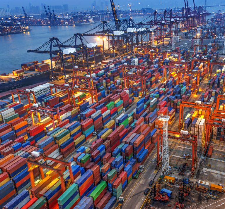 An aerial view of a busy shipping port at dusk, filled with colorful stacked shipping containers, large cranes, and cargo ships docked by the water, with city buildings visible in the background.