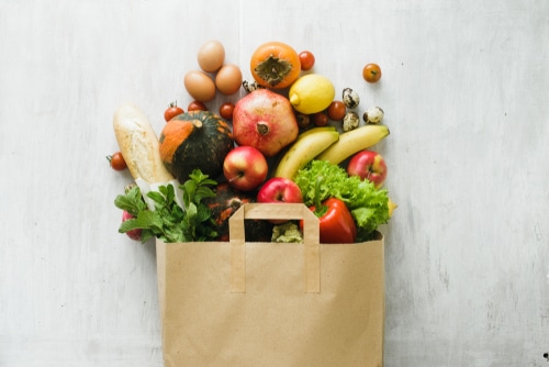A paper bag filled with fresh groceries, including apples, bananas, pomegranate, eggs, tomatoes, lettuce, a loaf of bread, and a variety of vegetables, against a light background.