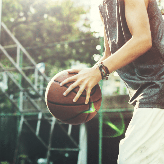 A person wearing a sleeveless shirt dribbles a basketball on an outdoor court, with sunlight filtering through trees in the background.