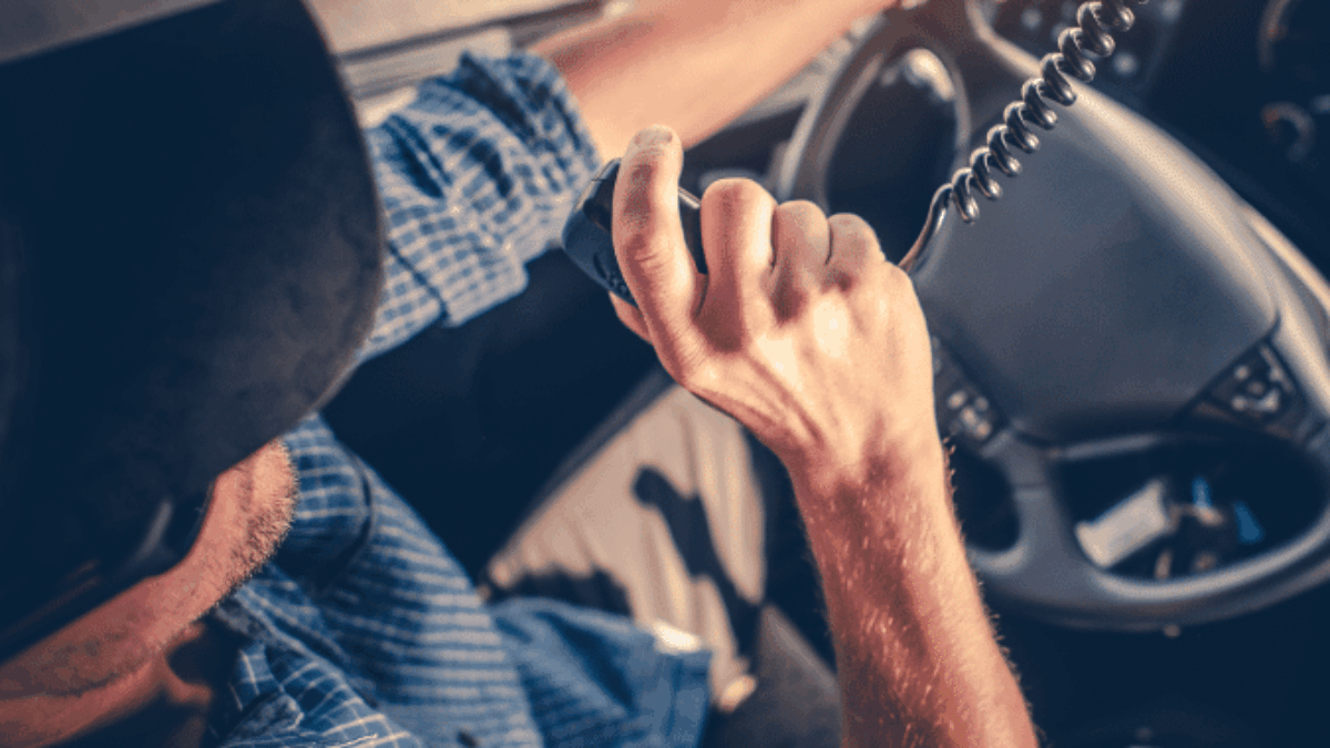 A person wearing a cap and a blue plaid shirt drives a vehicle while holding and speaking into a CB radio microphone, with sunlight shining through the window.