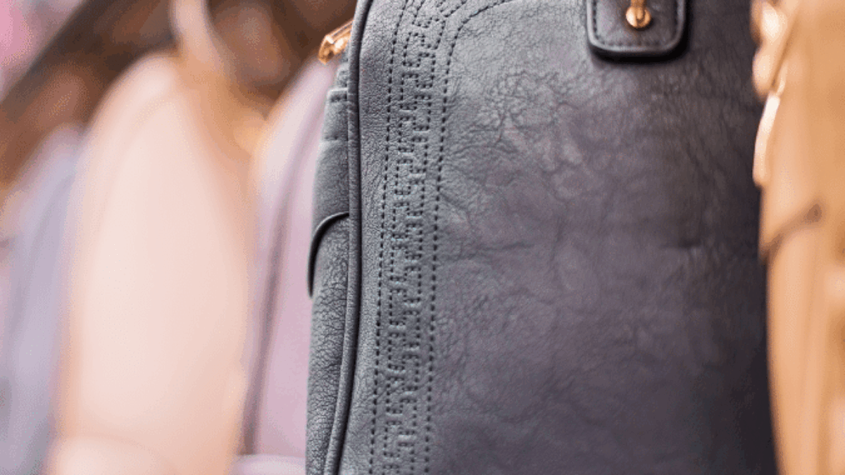 A close-up of a black leather handbag with gold hardware on a store shelf, surrounded by other handbags in various pastel colors.