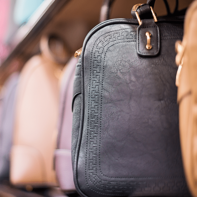 A close-up of a black leather handbag with gold hardware on a store shelf, surrounded by other handbags in various pastel colors.