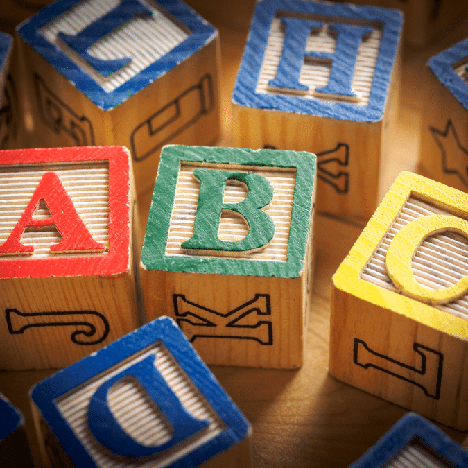 Colorful wooden alphabet blocks are arranged closely together, with the letters A, B, and D most prominently visible on the blocks faces. The blocks have textured surfaces and painted borders in red, green, blue, and yellow.