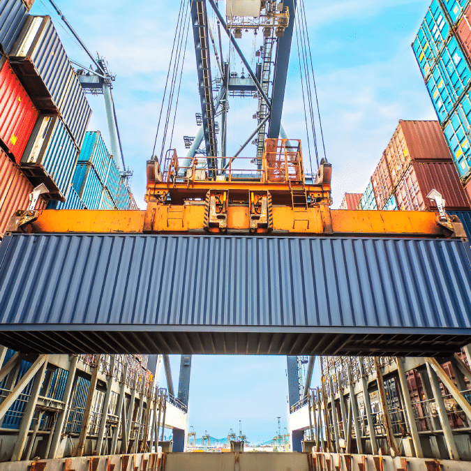 A large crane lifts a shipping container at a busy port, surrounded by stacked colorful containers under a blue sky.