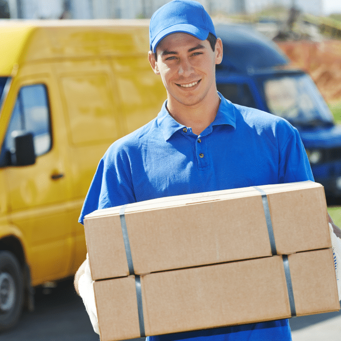 A smiling delivery person in a blue uniform and cap holds two cardboard boxes. A yellow van and a blue van are parked in the background.