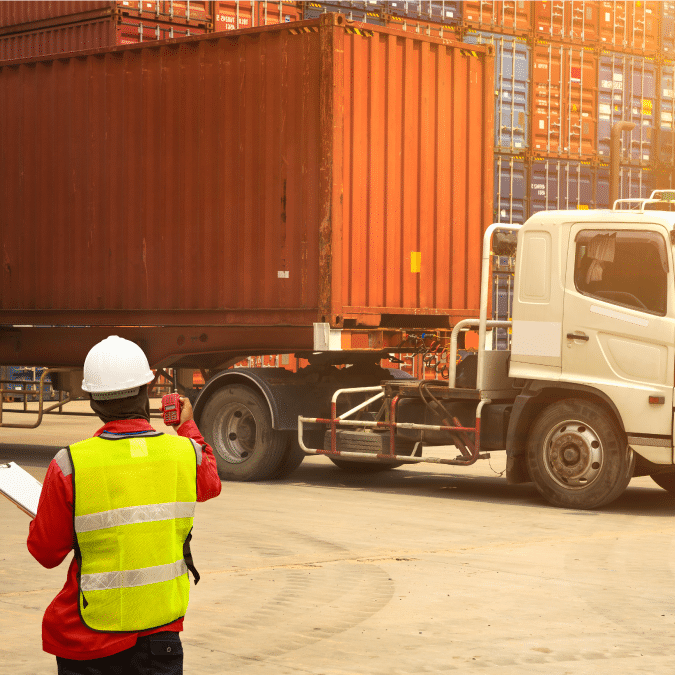 A worker in a reflective vest and hard hat inspects a large shipping container on a truck at a busy shipping yard with stacked containers in the background.