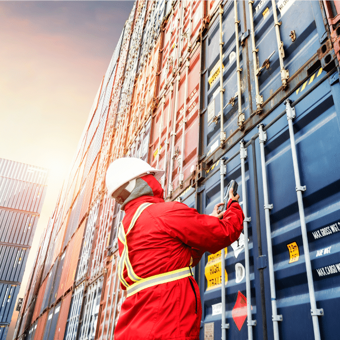 A worker in a red uniform and white hard hat inspects or secures stacked shipping containers outdoors, with sunlight shining in the background.
