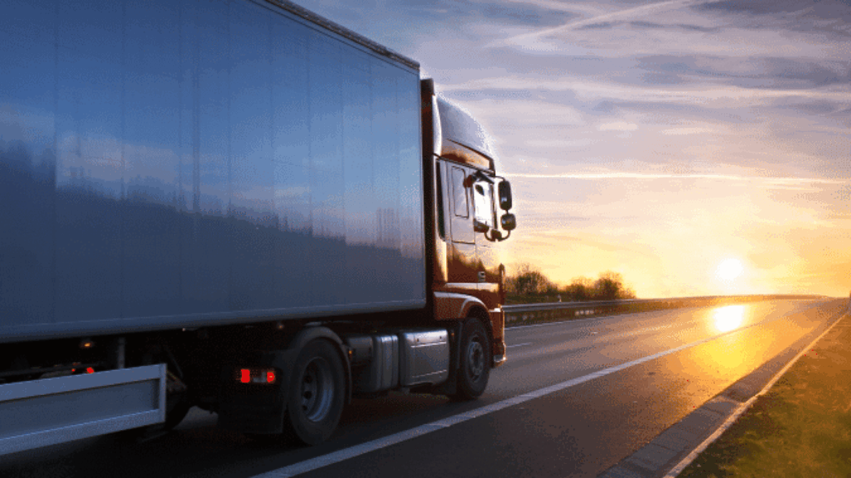 A large semi-truck drives on an empty highway at sunset, with the sky displaying orange and blue hues and the sun low on the horizon.