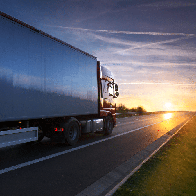 A large semi-truck drives on an empty highway at sunset, with the sky displaying orange and blue hues and the sun low on the horizon.