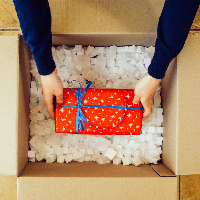 Person placing a red, star-patterned gift wrapped with a blue ribbon into a cardboard box filled with white packing peanuts, viewed from above.