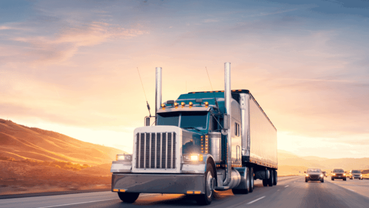 A large blue semi-truck drives down a highway at sunset, with a trailer attached and other cars visible in the adjacent lanes under a vibrant sky.