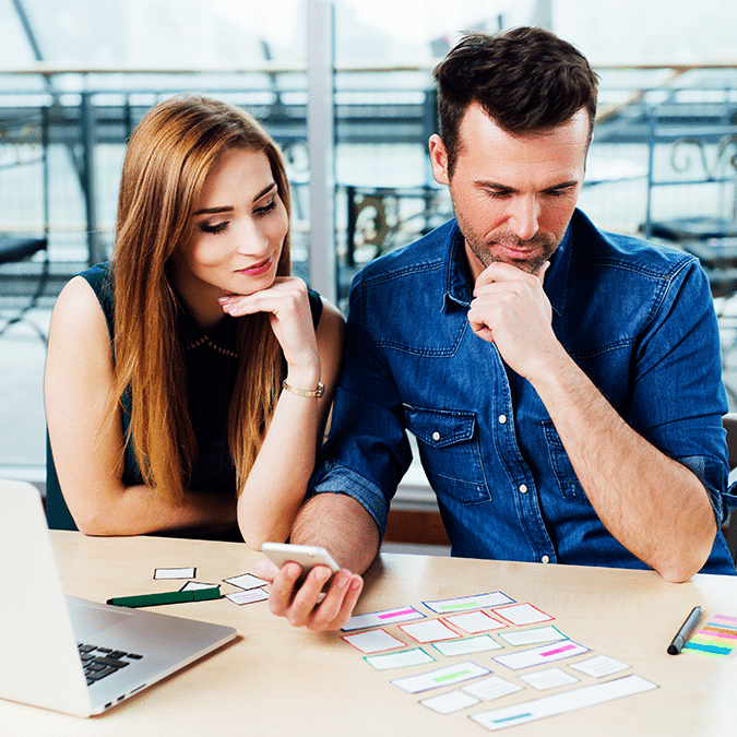A man and woman sit at a desk, thoughtfully looking at a smartphone and colorful cards arranged on the table. A laptop and pens are nearby, suggesting a collaborative planning or brainstorming session.