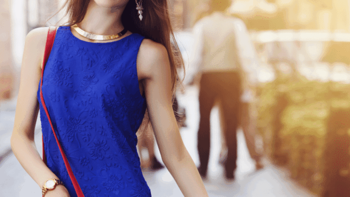 A stylish woman wearing a red hat, blue sleeveless top, white pants, and carrying a red purse walks down a sunlit city street, smiling confidently.