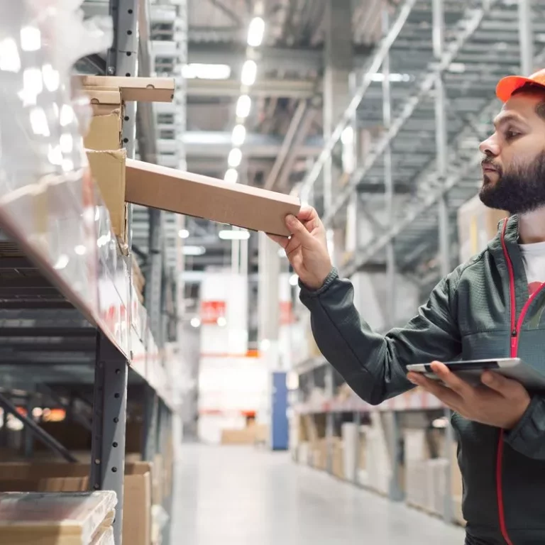 A man wearing an orange hard hat and a green jacket stands in a warehouse, holding a tablet and placing a box on a shelf. The warehouse has tall shelves stocked with various items.