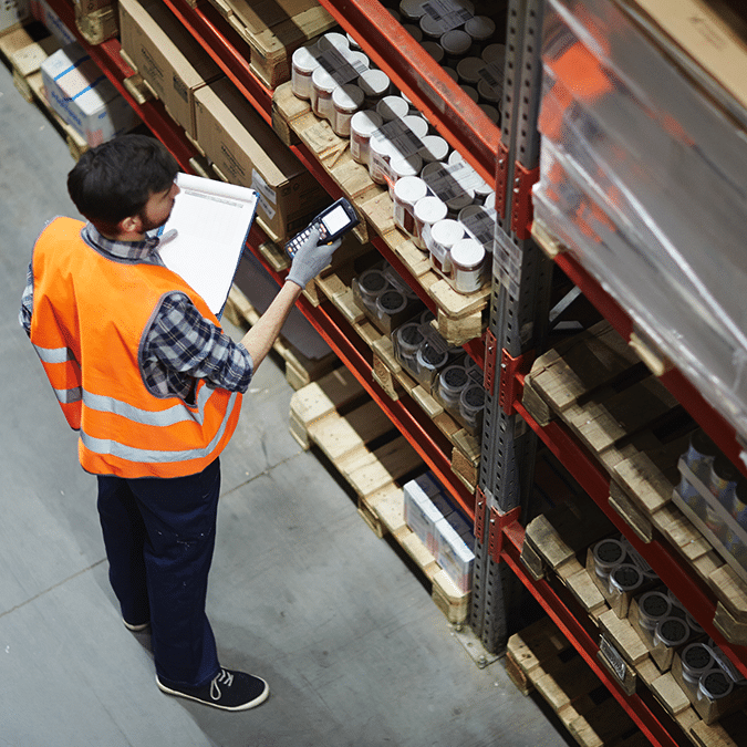 A warehouse worker wearing an orange safety vest and gloves uses a handheld scanner to check inventory on shelves, holding a clipboard and standing near stacks of boxed goods.