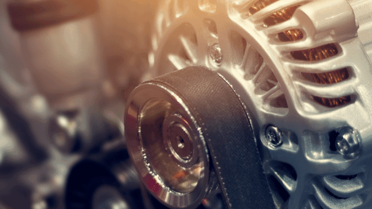 Close-up of a car engines alternator with a drive belt wrapped around a metallic pulley, surrounded by other engine components under warm lighting.