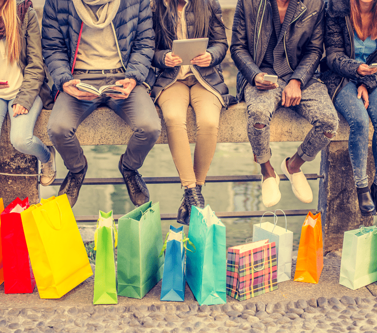 Five people sit on a stone bench outdoors, each with shopping bags of various colors and patterns placed in front of them. They are dressed in casual winter clothes and appear to be using their phones or reading.