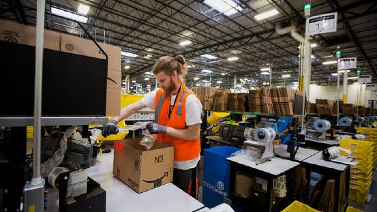 A worker wearing an orange safety vest packs a cardboard box at a workstation in a large warehouse filled with shelves, cardboard boxes, and packing materials.