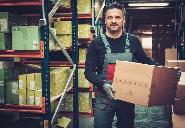 A warehouse worker in overalls and gloves holds a cardboard box, standing among shelves filled with packages and parcels. The storage area is organized and well-lit.
