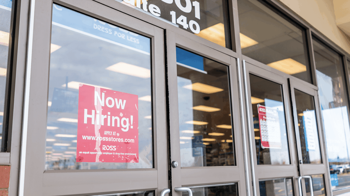 A glass storefront door displays a red “Now Hiring!” sign with application details for Ross. The store’s interior is visible through the windows, showing fluorescent lights and merchandise.