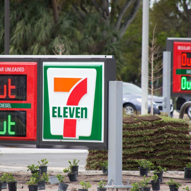 A 7-Eleven gas station sign displays OUT for both regular unleaded and diesel fuel, indicating they are unavailable. The same message appears on a second sign in the background. Small plants line the area.
