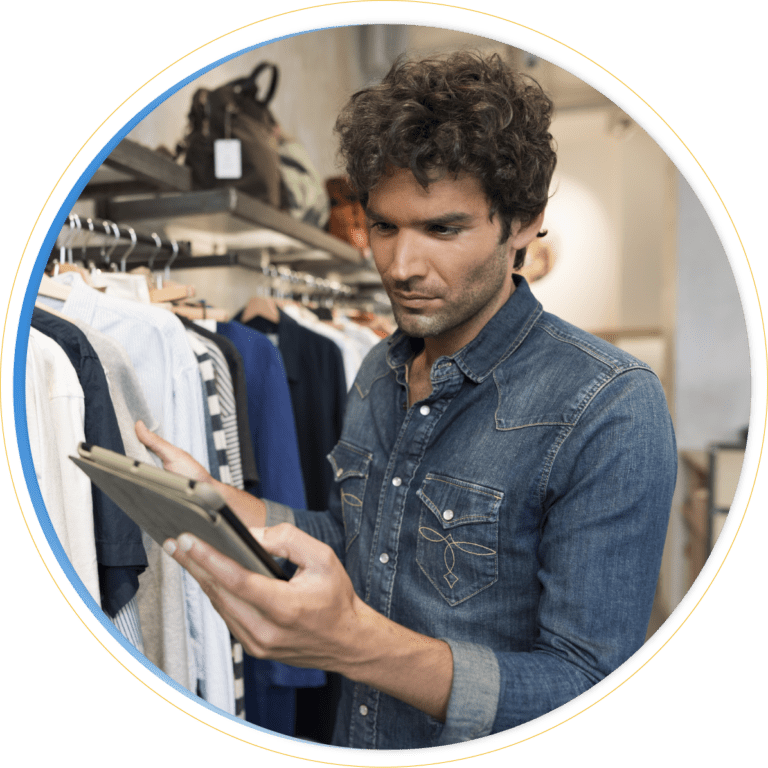 A man wearing a denim shirt stands in a clothing store, using a tablet to review shirts on a rack as part of the store’s merchandising strategy.