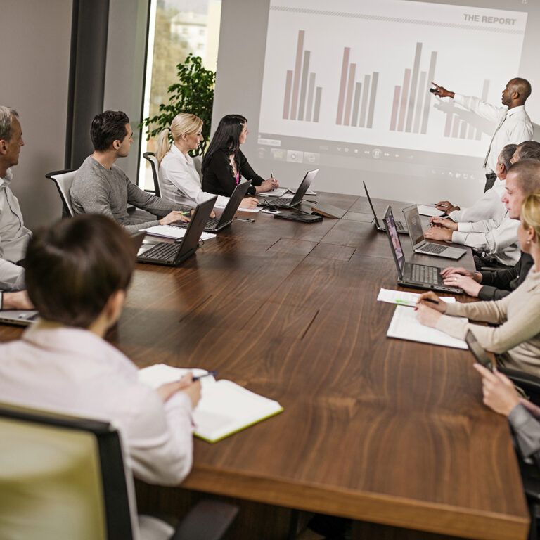 A group of people in business attire sit around a conference table with laptops and notepads, watching a presenter point to a bar chart on a projected screen during a meeting about the Microsoft Dynamics AX upgrade.